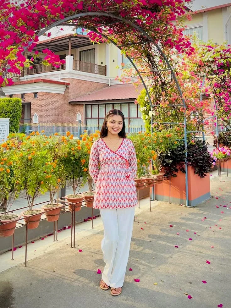 A photorealistic full-length portrait of a smiling young South Asian woman standing outdoors on a paved pathway beneath a blooming metal