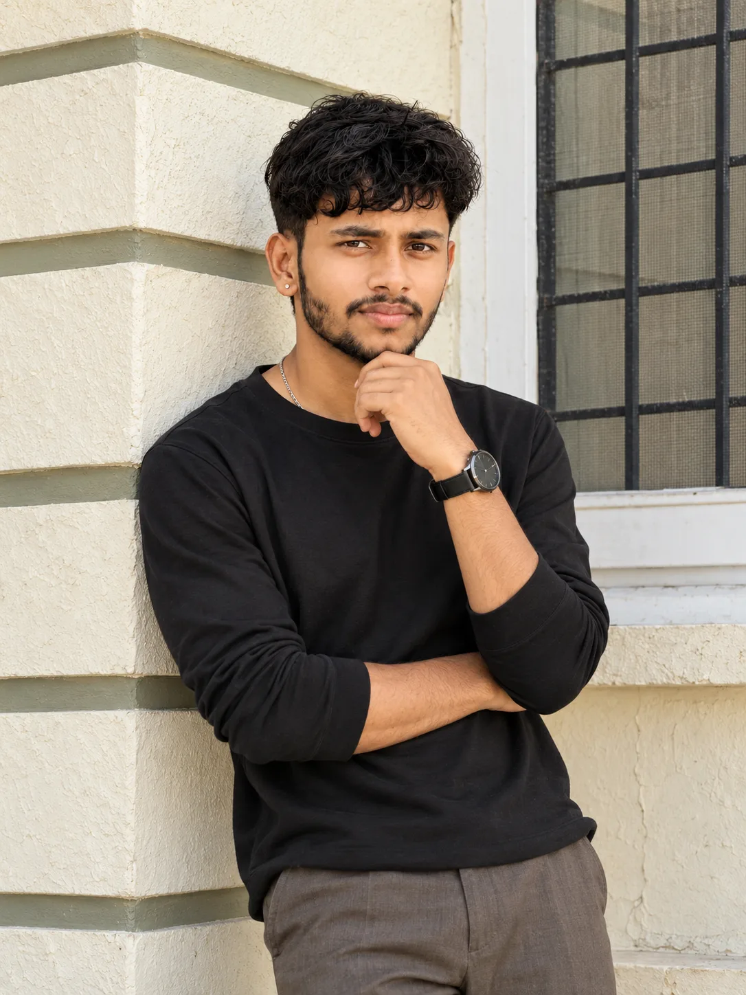 Ultra-realistic professional DSLR photograph of a slim young South Asian man standing casually against the corner of a pale cream textured